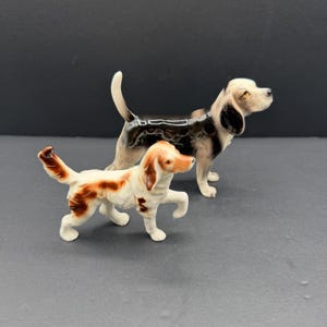 May include: Two ceramic dog figurines. One is a standing black, white, and tan hound with its tail raised. The second is a smaller, white and brown spotted dog in a walking pose with one front paw lifted, featuring a white tail with brown tips.