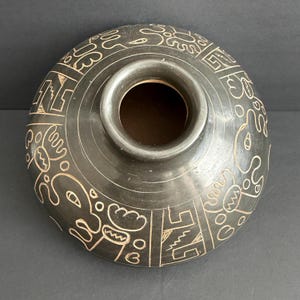 May include: A dark brown ceramic vessel with a wide opening and a rounded body. The surface is decorated with etched, cream-coloured abstract designs and geometric patterns. The vessel has a smooth, matte finish.