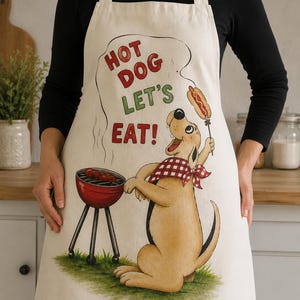 Könnte beinhalten: Cremefarbene Schürze mit einem Cartoon-Hund, der Hotdogs grillt. Der Hund trägt ein rot-weiß kariertes Halstuch und hält einen Hotdog am Spieß. Der Text über dem Hund lautet "HOT DOG LET'S EAT!" Ein roter Grill mit Hotdogs ist ebenfalls abgebildet.