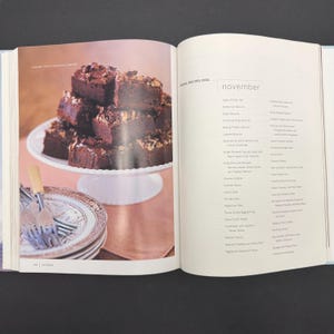 May include: An open cookbook displays a photo of stacked brownies on a white cake stand. The brownies are topped with nuts and chocolate drizzle. Beside the brownies are decorative plates and silverware. The opposite page lists recipes for November.