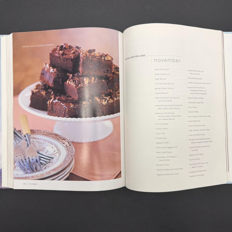 May include: An open cookbook displays a photo of stacked brownies on a white cake stand. The brownies are topped with nuts and chocolate drizzle. Beside the brownies are decorative plates and silverware. The opposite page lists recipes for November.