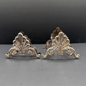 May include: A pair of silver-toned metal napkin holders featuring an ornate, openwork shell-like design with scrolling flourishes and small circular floral accents at the base. The structures are symmetrical with a curved, fan-shaped top edge.