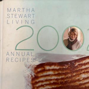 May include: A cookbook cover featuring the text "MARTHA STEWART LIVING" and "200" in large numbers. The words "ANNUAL RECIPES" are also visible. A portrait of Martha Stewart is in a circular frame. A layered dessert is in the foreground.
