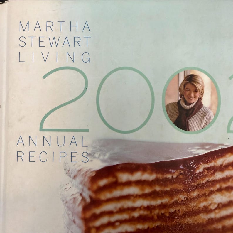 May include: A cookbook cover featuring the text "MARTHA STEWART LIVING" and "200" in large numbers. The words "ANNUAL RECIPES" are also visible. A portrait of Martha Stewart is in a circular frame. A layered dessert is in the foreground.