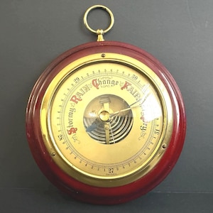 May include: A vintage barometer with a red wooden frame and a gold-coloured face. The face features a needle pointing to "Stormy," "Change," and "Fair" indicators. The barometer has a hanging ring at the top and is marked "Germany."