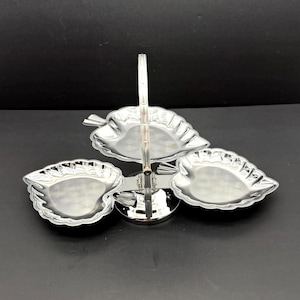 Op de afbeelding: Een zilverkleurige serveerschaal met drie niveaus. De schaal heeft een handvat en drie bladvormige schalen. De schalen zijn in een gelaagd ontwerp gerangschikt, met één schaal bovenop en twee eronder. De schaal is gemaakt van glanzend metaal.