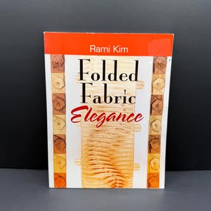 May include: A rectangular book titled Folded Fabric Elegance by Rami Kim. The cover features a central vertical panel of textured, light-colored fabric with a pleated design, flanked by two columns of small, square, folded fabric squares in shades of brown.