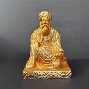 May include: A gold and silver colored figurine of a bearded figure wearing a turban and robes, seated in a cross-legged position on a rectangular base with a wavy pattern. The figure has one hand raised in a gesture and rests on a flat surface.