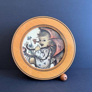 Puede incluir: Una caja de música de madera vintage con un marco redondo. La imagen interior presenta a un niño con una capucha roja, sosteniendo flores. La caja tiene un pequeño botón de madera en la parte inferior. El marco es marrón claro.