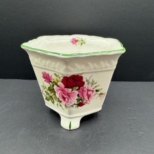 May include: A cream-colored ceramic planter with a square, flared rim and three feet. The exterior features a floral pattern of deep red and light pink roses with green leaves. A thin green line accents the top edge, and a small rose is painted inside.