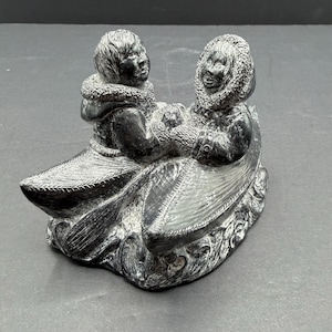May include: A dark grey stone sculpture depicting two figures seated in a canoe. The figures wear hooded parkas with textured trim and hold a paddle together. The base of the sculpture features carved details representing water waves.