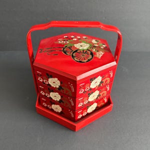 Op de afbeelding: Een rode, gelaagde bento-box in Japanse stijl met een handvat. De doos is versierd met witte bloemmotieven en een geschilderde scène van een kar met bloemen. De doos is gemaakt van gelakt hout.
