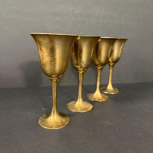 Puede incluir: Cuatro copas doradas vintage con un cuenco en forma de tulipán y un tallo esbelto. Las copas están dispuestas en fila, mostrando su elegante diseño y pátina envejecida. Las copas son de metal.