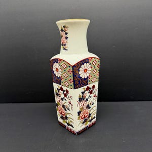 May include: A white ceramic vase with a square base and a flared top. The vase features floral and geometric patterns in red, blue, and gold. The design includes stylized flowers and intricate border details, creating a decorative piece.