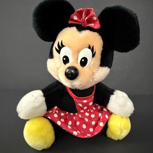 May include: A plush Minnie Mouse doll with black ears, a red bow with white polka dots, and a red and white polka dot dress. The doll has a black nose, large eyes, and yellow feet.