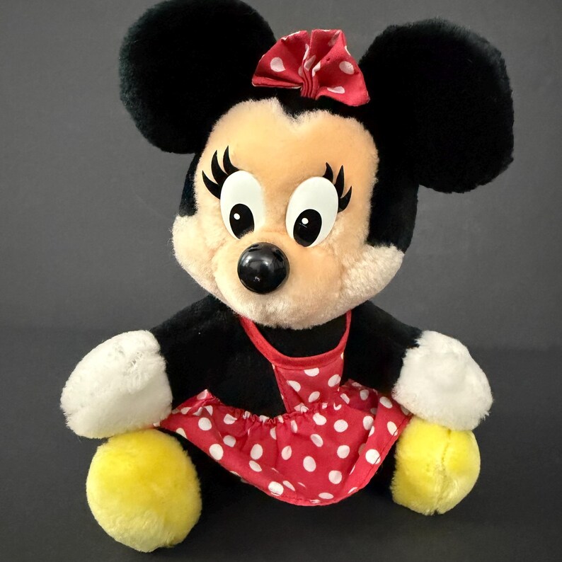 May include: A plush Minnie Mouse doll with black ears, a red bow with white polka dots, and a red and white polka dot dress. The doll has a black nose, large eyes, and yellow feet.
