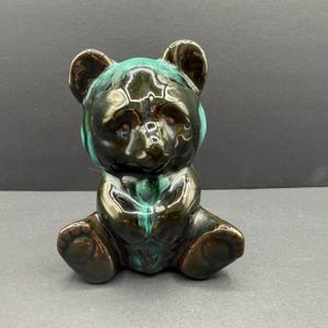 Könnte beinhalten: Eine Keramik-Bärenfigur in sitzender Position. Der Bär hat eine glänzende Oberfläche mit einer Farbpalette aus Braun, Schwarz und Türkis. Die Ohren des Bären sind abgerundet und seine Pfoten sind sichtbar. Die Figur ist ein dekorativer Gegenstand.