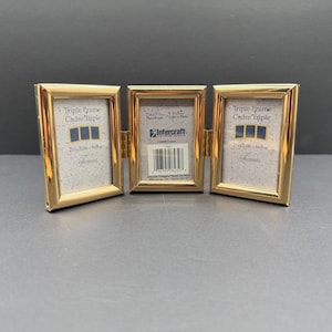 May include: A gold-colored triple photo frame with three hinged sections. Each frame is labeled "Triple Frame Cadre Triple" with dimensions of 2 3/8 x 3 1/2 inches and 6 x 8 inches. The center frame has a barcode and the Intercraft logo.