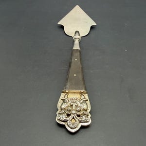 May include: A vintage cheese knife with a silver blade and a dark wooden handle. The handle features a decorative silver end with an ornate face design. The knife is approximately 9 inches long.