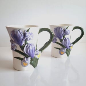 May include: Two white ceramic mugs featuring a sculpted iris flower design. The irises are purple with yellow details, and the stems and handles are green. The mugs have a distinctive, angular form.