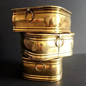 May include: Three stacked, rectangular brass boxes with rounded edges and ring handles. The boxes are highly reflective, with a polished, golden finish. The boxes are arranged in a vertical stack, with each box slightly smaller than the one below it.