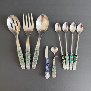 Könnte beinhalten: Eine Sammlung handgefertigter Silberwaren mit dekorativen Griffen. Das Set umfasst einen Schöpflöffel, eine Gabel, einen Löffel, ein Buttermesser und einen kleinen Löffel sowie fünf langstielige Löffel. Die Griffe sind mit Draht umwickelt und mit grünen und blauen Perlen verziert.