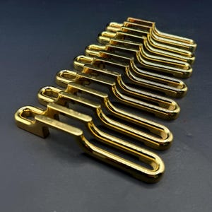 May include: A collection of eight identical gold-toned metal sliders arranged in a parallel row. Each slider features a smooth, reflective finish with a rectangular loop design, characterized by a raised bridge section and an elongated opening.