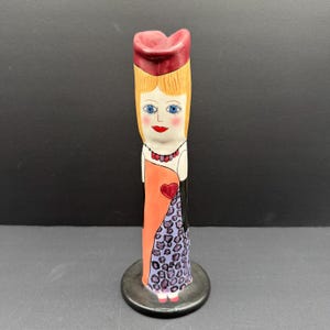 May include: A tall ceramic vase shaped like a stylised figure wearing a red hat, a peach-coloured dress with a red heart, and a purple leopard-print skirt. The figure has blonde hair, blue eyes, red lips, and a beaded necklace on a white base.