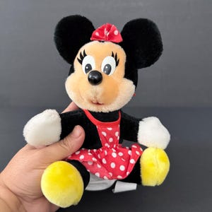 May include: A Minnie Mouse plush toy. The plush has black fur, a red and white polka dot dress, and a red bow. The toy has yellow feet and white gloves. The plush is being held by a hand.