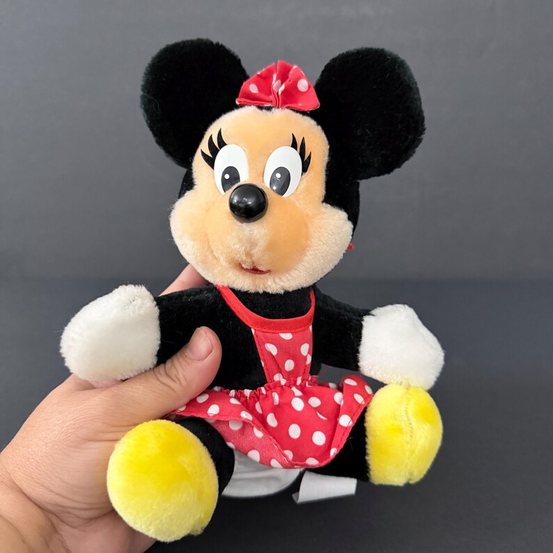 May include: A Minnie Mouse plush toy. The plush has black fur, a red and white polka dot dress, and a red bow. The toy has yellow feet and white gloves. The plush is being held by a hand.