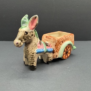 Puede incluir: Una figura de cerámica de un burro tirando de un carro, con pelaje moteado marrón y bronceado, orejas forradas de rosa y detalles verdes. El carro tiene un exterior naranja texturizado, un arco de rueda verde claro y una rueda de radios marrón.