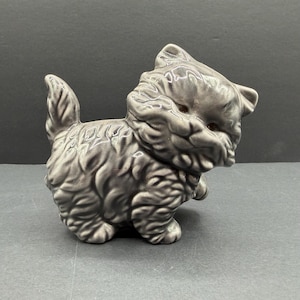 May include: A glossy, grey ceramic figurine of a fluffy kitten standing with its tail raised. The sculpture features detailed, textured fur patterns across its body, pointed ears, and dark, inset eyes, capturing a playful, alert posture.