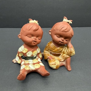 May include: Two terracotta-colored figurines of seated figures with closed eyes. One wears a patterned kimono with white and yellow circles, the other a gold-colored robe. Both have small topknots.