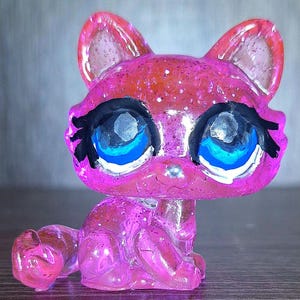 May include: A small, translucent pink cat figurine with a glittery finish. The figure features large, stylized eyes with bright blue irises, white highlights, and thick black eyelashes. The cat is in a seated position with its tail curled forward.
