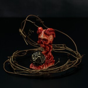 May include: A red and white skull-shaped figurine with horns and a tentacle-like base, holding a black and gold polyhedron die. The figurine is surrounded by a circular arrangement of thorny branches, all set against a dark background.