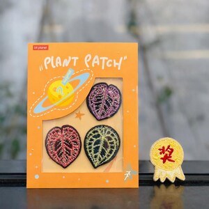 May include: An orange card with the text "PLANT PATCH" contains three leaf-shaped embroidered patches in red, purple, and green. A small yellow patch with red text is also visible. The card has a planet graphic.