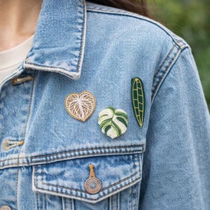Pode incluir: Jaqueta jeans azul clara adornada com três patches bordados com tema de plantas. Os patches incluem uma folha em forma de coração, um filodendro de folha dividida e uma folha longa verde. A jaqueta tem um design clássico com bolsos abotoados e gola.