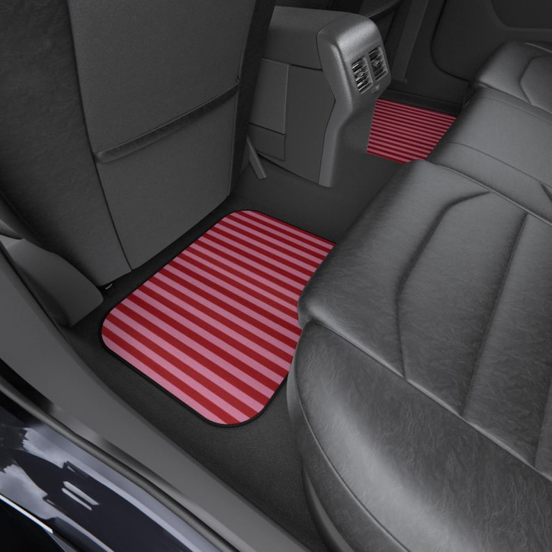 May include: A set of rectangular car floor mats featuring a repeating pattern of alternating dark red and light pink horizontal stripes. The mats are positioned on the floor of a vehicle interior with black leather seats and a center console.