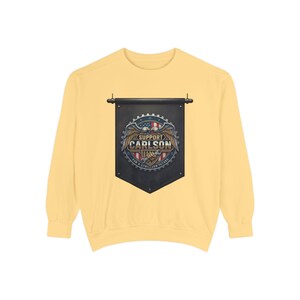May include: A light yellow sweatshirt featuring a black banner with the text "SUPPORT CARLSON" and an eagle design. The banner has a circular emblem with stars and stripes. The sweatshirt has long sleeves and a crew neck.