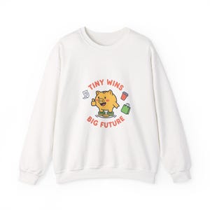 Könnte beinhalten: Weißes Sweatshirt mit einer Grafik eines lächelnden gelben Sparschweins, das einen Daumen hoch gibt, umgeben von Münzen, einer Einkaufstasche, einem Kaffeebecher und einem Dokument. Der Text lautet "Tiny Wins Big Future".