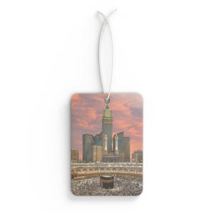 May include: Rectangular air freshener featuring a cityscape with tall buildings and a large crowd surrounding a central structure. The sky is a blend of orange and pink hues. A white string is attached for hanging.