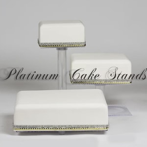 May include: Three-tiered white cake stands with a square design. Each stand has a decorative trim of silver and gold rhinestones. The stands are supported by clear acrylic columns. The text "Platinum Cake Stands" is visible.
