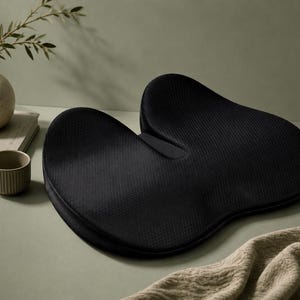 May include: A black ergonomic seat cushion with a contoured shape. The cushion is made of a textured fabric. A small ceramic cup, a vase with greenery, and a book are also in the image. The cushion is designed for support.