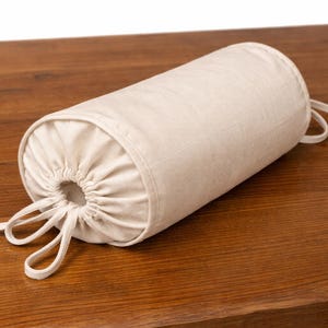 May include: A cylindrical bolster cushion in a neutral beige fabric, with gathered ends and tie closures. The cushion is on a wooden surface, highlighting its shape and texture. This decorative cushion is suitable for home decor.