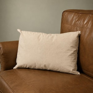 May include: A rectangular throw pillow with a solid light beige fabric cover. The pillow has a smooth, plain texture and is positioned horizontally, resting against the surface of a brown leather armchair with visible stitched seams.