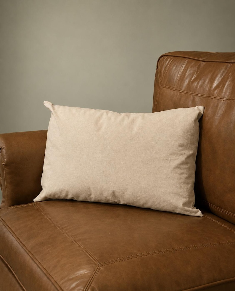 May include: A rectangular throw pillow with a solid light beige fabric cover. The pillow has a smooth, plain texture and is positioned horizontally, resting against the surface of a brown leather armchair with visible stitched seams.
