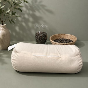 May include: A beige cylindrical yoga bolster with a white zip, set on a sage green surface. A clear glass vase filled with dark peppercorns and a small woven basket with peppercorns are in the background. A beige vase with a green leafy branch is also in the background.