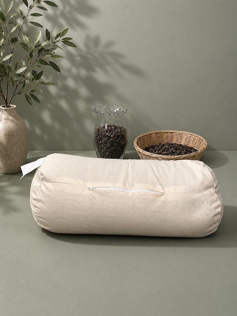 May include: A beige cylindrical yoga bolster with a white zip, set on a sage green surface. A clear glass vase filled with dark peppercorns and a small woven basket with peppercorns are in the background. A beige vase with a green leafy branch is also in the background.