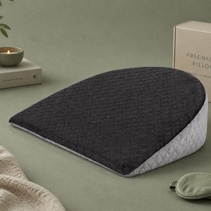 May include: A dark gray quilted pregnancy pillow with a light gray side panel. The pillow is on a green surface with a box that reads "PREGNANCY PILLOW" and a sleep mask.