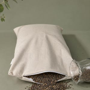 May include: A rectangular beige fabric pillow with a white zipper closure partially open at the bottom. Dark brown buckwheat hulls are spilling out from the opening onto the surface, and a clear glass vessel is positioned next to the pillow.